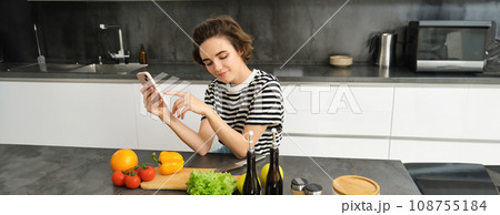 Portrait of cute brunette woman in the kitchen, cooking, searching recipe on smartphone, taking pictures for her food blog, cooking meal from vegetables, vegan salad Portrait of cute brunette woman in the kitchen, cooking, searching recipe on smartphone, taking pictures for her food blog, cooking meal from vegetables, vegan salad 108755184
