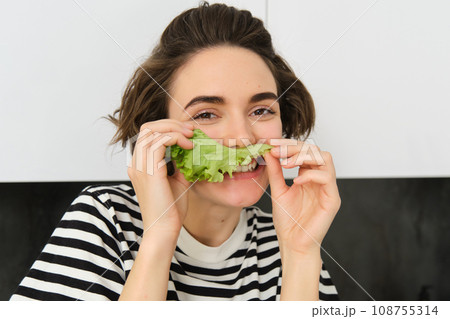 Close up portrait of young woman, vegetarian girl, likes eating vegetables, posing with lettuce leaf and smiling, posing in the kitchen. Concept of healthy food and diet 108755314