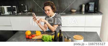 Portrait of young woman with smartphone, sitting in the kitchen and cooking salad, searching for healthy recipe online, has vegetables and chopping board on the table 108755327