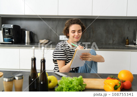 Smiling brunette woman in kitchen, writing down meal list for week, reading recipe, cooking salad, vegetable meal at home Smiling brunette woman in kitchen, writing down meal list for week, reading recipe, cooking salad, vegetable meal at home 108755341