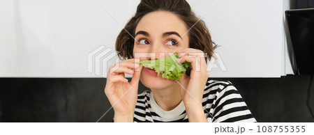 Close up of funny cute woman, vegetarian eating lettuce leaf and smiling, concept of healthy diet, girl likes vegetables, standing in the kitchen 108755355