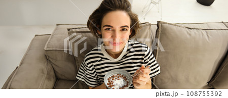 Close up of cute young female model, eating cereals with milk, enjoys her breakfast on sofa in living room, smiling and looking happy Close up of cute young female model, eating cereals with milk, enjoys her breakfast on sofa in living room, smiling and looking happy 108755392