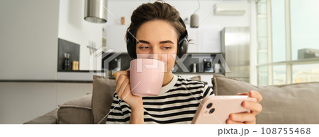 Close up of woman watching video with interest, looking at smartphone screen, wearing headphones and drinking tea from pink cup, sitting on sofa in living room at home 108755468