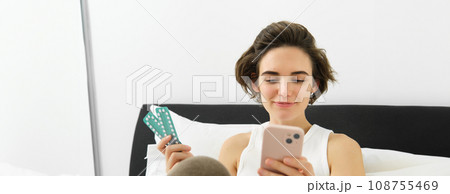 Portrait of woman in bed, holding mobile phone and pills, taking medication, searching for dosage and instruction for tablets, resting at home Portrait of woman in bed, holding mobile phone and pills, taking medication, searching for dosage and instruction for tablets, resting at home 108755469