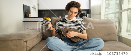Lifestyle and hobbies concept. Young smiling woman on sofa, playing ukulele, singing and learning strumming pattern for favourite song 108755470