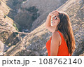 Young attractive brunette woman sitting on the background of the mountains, enjoying the sunshine. Concept of traveling, hiking, vacation and leisure Young attractive brunette woman sitting on the background of the mountains, enjoying the sunshine. Concept of traveling, hiking, vacation and leisure 108762140