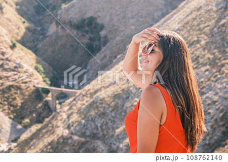 Young attractive brunette woman sitting on the background of the mountains, enjoying the sunshine. Concept of traveling, hiking, vacation and leisure 108762140