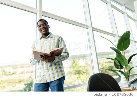 African man standing near the window in room and reading a book 108765036