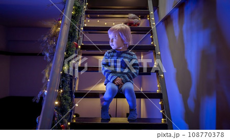 Little boy sitting on stairs decorated for Christmas or New Year and looking at police lights and siren. Concept of Crime on winter holidays, theft, burglar in house and protection Little boy sitting on stairs decorated for Christmas or New Year and looking at police lights and siren. Concept of Crime on winter holidays, theft, burglar in house and protection 108770378