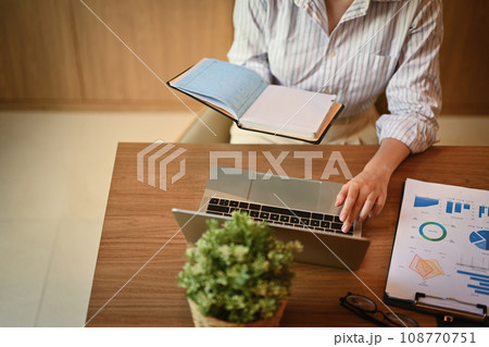 Cropped shot of female manager holding her personal daily planner and using laptop at desk Cropped shot of female manager holding her personal daily planner and using laptop at desk 108770751