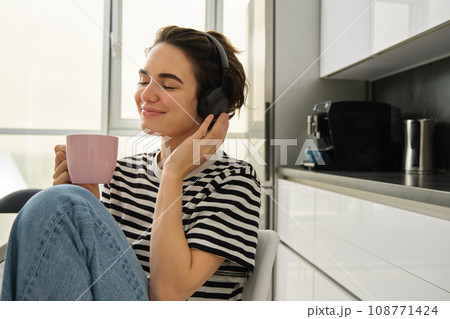 Lifestyle and music concept. Happy smiling woman with cup of tea, sits in kitchen and touches headphones on her head, listens to music with pleased relaxed face 108771424