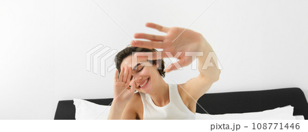 Portrait of sleepy, happy young woman, wakes up in her bed, stretching out hands, hiding face from camera, smiling and laughing Portrait of sleepy, happy young woman, wakes up in her bed, stretching out hands, hiding face from camera, smiling and laughing 108771446