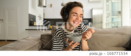 Close up of happy beautiful woman, laughing and smiling, watching tv in wireless headphones, holding bowl of cereals, having breakfast in living room on sofa 108771534