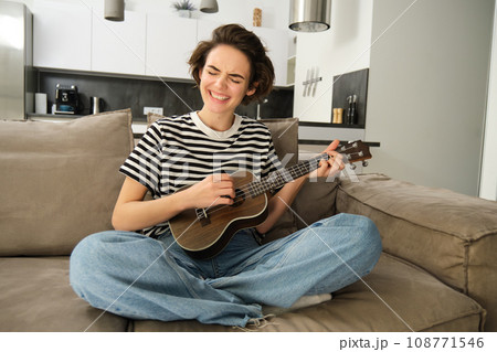Young woman playing ukulele with passion, singing song, strumming strings, sitting on sofa in living room at home 108771546