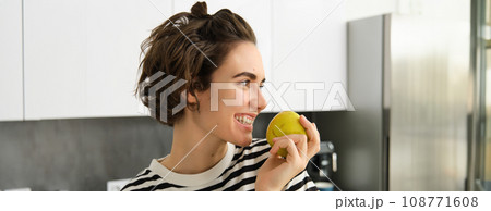 Close up portrait of cute young modern woman, eating green apple and looking aside, smiling while biting a fruit, having a snack in the kitchen Close up portrait of cute young modern woman, eating green apple and looking aside, smiling while biting a fruit, having a snack in the kitchen 108771608