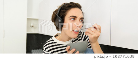 Cute young woman in headphones, eating with spoon from bowl, having cereals for breakfast, sitting in kitchen in casual striped t-shirt Cute young woman in headphones, eating with spoon from bowl, having cereals for breakfast, sitting in kitchen in casual striped t-shirt 108771639