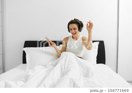 Portrait of happy woman, wakes up and dancing in wireless headphones, listens to music and sings, holds smartphone 108771669