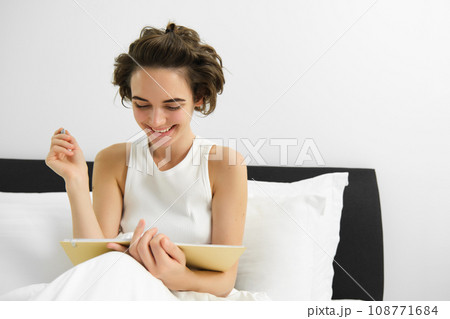 Portrait of woman sitting in bed with journal, writing in diary with pen, reading notebook, studying or doing homework from her bedroom Portrait of woman sitting in bed with journal, writing in diary with pen, reading notebook, studying or doing homework from her bedroom 108771684
