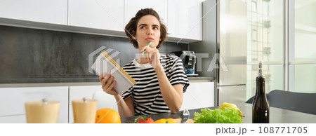 Portrait of thinking woman with notebook, cooking, writing down recipe ingredients, deciding on a meal for dinner, sitting near vegetables and chopping board in kitchen Portrait of thinking woman with notebook, cooking, writing down recipe ingredients, deciding on a meal for dinner, sitting near vegetables and chopping board in kitchen 108771705