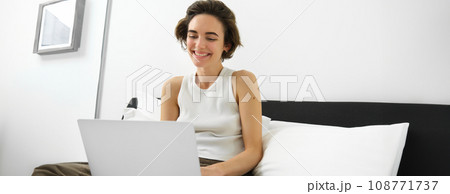 Image of smiling, happy young woman in bedroom, sitting on bed with laptop, laughing and looking at computer screen, working from home, freelancing 108771737