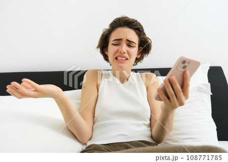 Portrait of woman with confused face, laying in bed with smartphone, chatting on mobile phone app, video call, shrugging shoulders at some nonsense, feeling upset by something she sees online 108771785