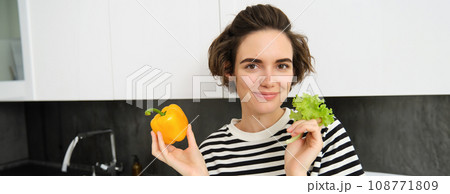 Close up of cute smiling woman, showing lettuce leaf and yellow pepper, cooking salad in the kitchen, likes eating vegetables as healthy snack Close up of cute smiling woman, showing lettuce leaf and yellow pepper, cooking salad in the kitchen, likes eating vegetables as healthy snack 108771809