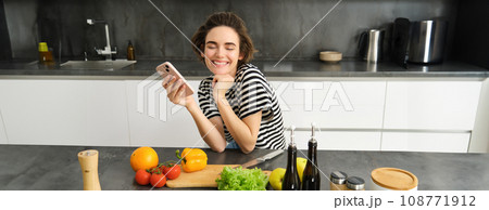 Happy smiling woman, sitting in the kitchen with smartphone, cooking salad, has chopping board, olive oil and vegetables on table 108771912