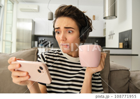 Portrait of woman with worried face expression, watching intense scene in tv show on mobile phone app, drinking tea, frowning and staring at smartphone, sitting on sofa in living room 108771955