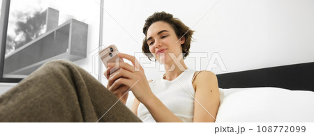 Portrait of young beautiful woman lying in bed, resting in bedroom, messaging, using mobile phone, holding smartphone and smiling, scrolling social media, spending comfort time at home 108772099