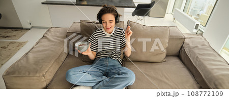 Portrait of stylish young woman listening to music in headphones, eating cereals with milk on sofa in living room, dancing while sitting and having breakfast, holding bowl and spoon 108772109