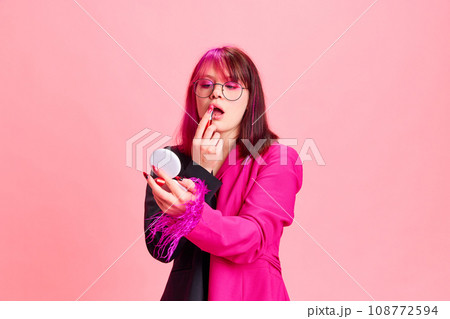 Young girl with pink hair and colorful jacket applying lipsticks against pink studio background 108772594