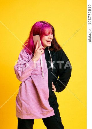 Smiling and laughing young girl with colorful hair and piercing talking on mobile phone against vivid yellow studio background 108772619