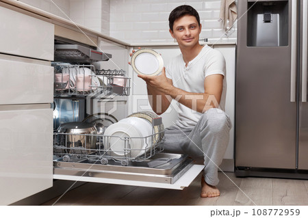 handsome positive man takes out clean dishes from the dishwasher in white modern kitchen 108772959