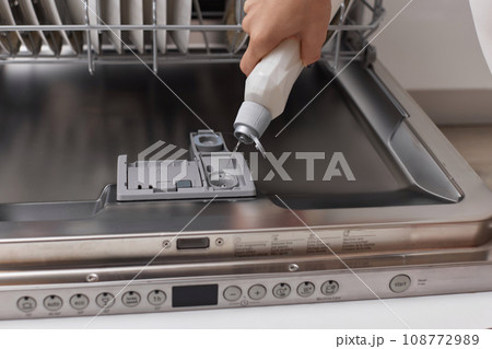 female hand pours rinse aid into the dishwasher compartment in modern white kitchen 108772989