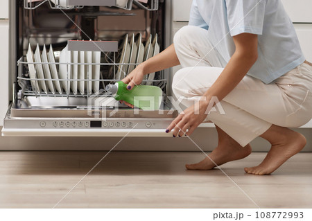 female hand pours rinse aid into the dishwasher compartment in modern white kitchen 108772993