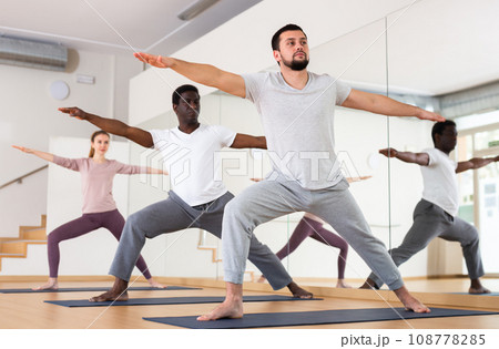 Bearded man standing in Virabhadrasana with group in yoga studio Bearded man standing in Virabhadrasana with group in yoga studio 108778285