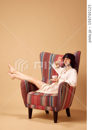 Young pretty lady sitting in armchair in silk bathrobe and mask of sleeping with cup of hot coffee or tea isolated beige studio background. Young pretty lady sitting in armchair in silk bathrobe and mask of sleeping with cup of hot coffee or tea isolated beige studio background. 108780125