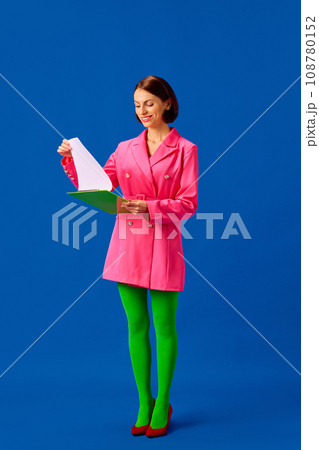Full length portrait of young woman, business lady dressed smart casual vivid look stands looking to folder and checking tasks isolated blue background. Full length portrait of young woman, business lady dressed smart casual vivid look stands looking to folder and checking tasks isolated blue background. 108780152