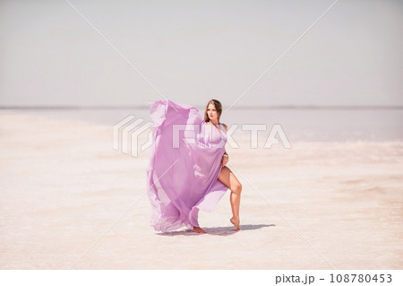 Woman pink salt lake. Against the backdrop of a pink salt lake, a woman in a long pink dress takes a leisurely stroll along the white, salty shore, capturing a wanderlust moment. 108780453