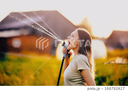 Watering vegetables from garden hose, young woman splashes water while fooling around and making faces. 108782627