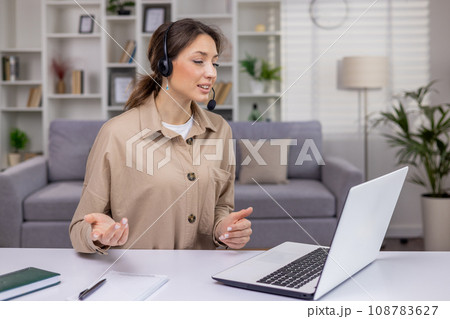Businesswoman in headset sitting at table in living room in front of laptop, communicating via video call, attractive woman working in call center, working from home. 108783627