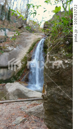 vertical image of a small waterfall in blurred focus visible from behind a rocky ledge, natural landscape for hiking and tourism in the mountains of the Republic of Adygea vertical image of a small waterfall in blurred focus visible from behind a rocky ledge, natural landscape for hiking and tourism in the mountains of the Republic of Adygea 108783722