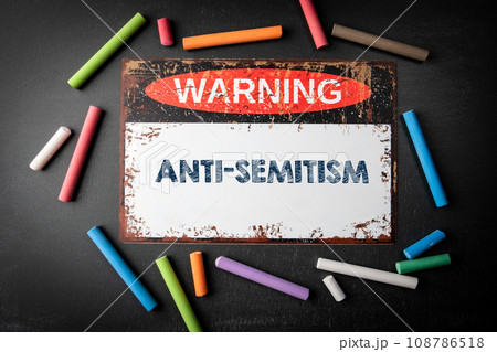 Anti-Semitism. Metal warning sign and colored pieces of chalk on a dark chalkboard background Anti-Semitism. Metal warning sign and colored pieces of chalk on a dark chalkboard background 108786518