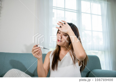 Sick Asian young woman sit under blanket on sofa she sneeze checking temp with digital thermometer and tissue paper in living room at home, Cold flu and migraine concept, medical healthcare 108789400