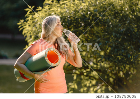Senior woman with yoga mat standing at park with bottle of water. Positive mature woman after fitness or yoga class in nature. Wellness and healthy lifestyle on retirement. 108790608