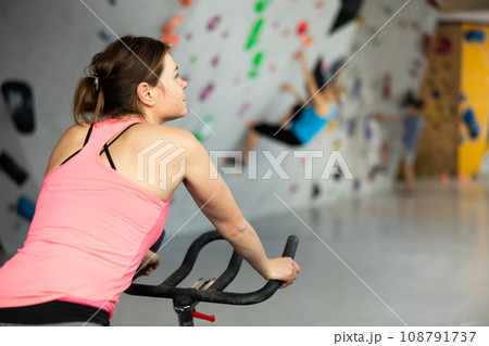 Sports girl on an exercise bike in climbing hall Sports girl on an exercise bike in climbing hall 108791737