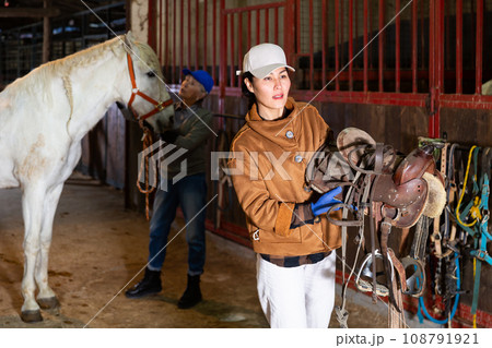Woman horse breeder carrying leather saddle Woman horse breeder carrying leather saddle 108791921
