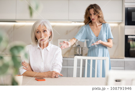 Adult woman quarreling with elderly woman in kitchen Adult woman quarreling with elderly woman in kitchen 108791946