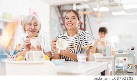 Cheerful young girl and older woman decorating ceramic mugs in pottery workshop 108792102