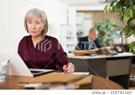 Concentrated elderly female office employee working with laptop Concentrated elderly female office employee working with laptop 108792160